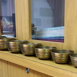 Puede incluir: Un conjunto de ocho cuencos cantores, hechos de latón, con diseños intrincados. Los cuencos están dispuestos en una fila sobre una superficie de madera.