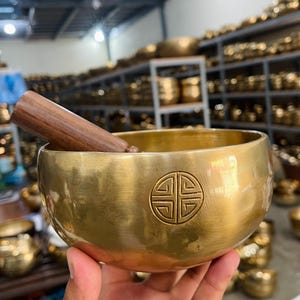 Könnte beinhalten: Eine goldene Klangschale mit einem Holzschlägel. Die Schale hat ein traditionelles Design mit einem kreisförmigen Muster in der Mitte. Die Schale wird von einer Hand gehalten.