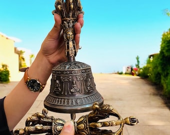 May include: A bronze-colored Buddhist bell with intricate carvings and a crown-like top is held up against a blue sky. Below the bell is a matching bronze vajra, a ritual object. The bell features detailed relief carvings of seated figures.