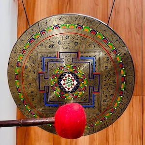 Op de afbeelding: Een grote, sierlijke gong met een gedetailleerd, goudkleurig ontwerp. De gong heeft ingewikkelde patronen en kleurrijke accenten van rood, blauw, groen en geel. Een rode hamer ligt voor de gong. De gong is opgehangen aan een zwart koord.