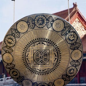 Op de afbeelding: Een grote, ronde gong met ingewikkelde gouden en zwarte patronen. De gong heeft gedetailleerde religieuze symbolen en tekst, opgehangen aan een touw. De achtergrond bevat een traditioneel gebouw met een rood dak.