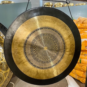 Op de afbeelding: Een grote, ronde gong met een zwarte buitenrand en een goudkleurig midden. Het midden heeft een ingewikkeld geometrisch patroon. De gong is opgehangen aan een zwart koord aan een metalen staaf.