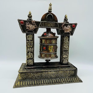 May include: A gold and copper prayer wheel with intricate carvings and red gemstones, mounted on a decorative stand with a traditional design. The wheel is inscribed with Tibetan script.