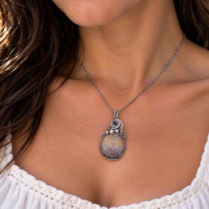 May include: A teardrop-shaped pendant necklace featuring a multi-colored, patterned stone encased in intricate, oxidized silver wire wrapping. The pendant hangs from a delicate silver chain, accented with small silver beads at the top.