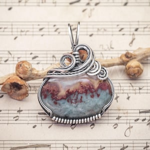 May include: A wire-wrapped pendant featuring a polished, oval-shaped stone with horizontal bands of muted blue, cream, and deep red. The stone is encased in silver-toned metal wire, which is coiled into decorative swirls at the top and base.