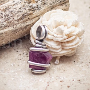 May include: A faceted, deep magenta gemstone pendant wrapped in silver wire. The pendant has a loop at the top for a chain. The background includes a piece of driftwood and a cream-colored flower.