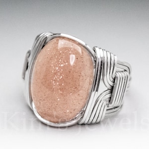 May include: A silver wire wrapped ring with a light pink oval gemstone. The gemstone has a sparkly, speckled texture.