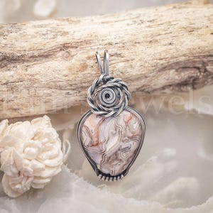 May include: A pendant featuring a teardrop-shaped stone with intricate white, tan, and brown swirling patterns, encased in a silver wire-wrapped frame. The top of the pendant is adorned with a decorative silver wire coil and a bail for hanging.