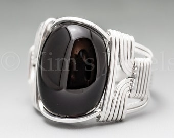 May include: A silver-toned ring featuring a large, oval-shaped, polished black stone. The stone is set within a wire-wrapped design, with the silver wire forming intricate patterns around the stone and band. The ring has a modern, handcrafted aesthetic.