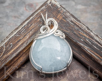 May include: A light blue aquamarine gemstone pendant wrapped in silver wire. The pendant is on a wooden surface.
