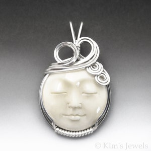 May include: A white ceramic pendant with a sculpted face, closed eyes, and a serene expression. The pendant is wrapped in silver wire with intricate swirls and loops.