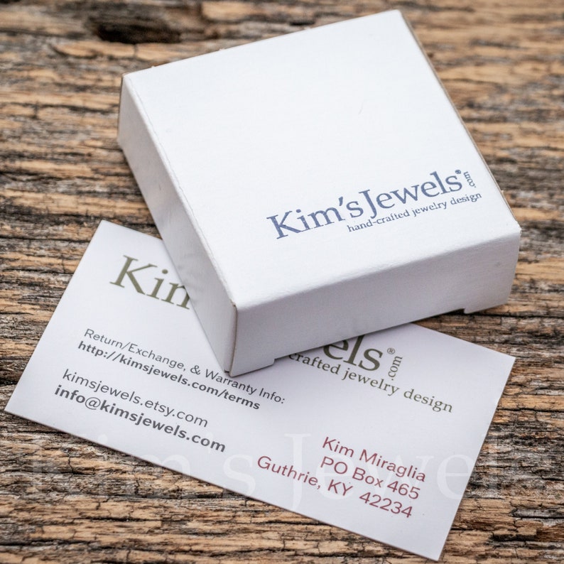 May include: A white box with the text "Kim's Jewels hand-crafted jewelry design" on it. A business card with the text "Kim's Jewels crafted jewelry design" and contact information is on a wooden surface.