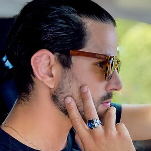 May include: A man with dark hair tied back in a bun is wearing brown tortoiseshell sunglasses with the brand name "DOMIZETTI" printed on the frame. He is wearing a blue shirt and a gold chain necklace. He is wearing a silver ring with a blue gemstone on his right hand.