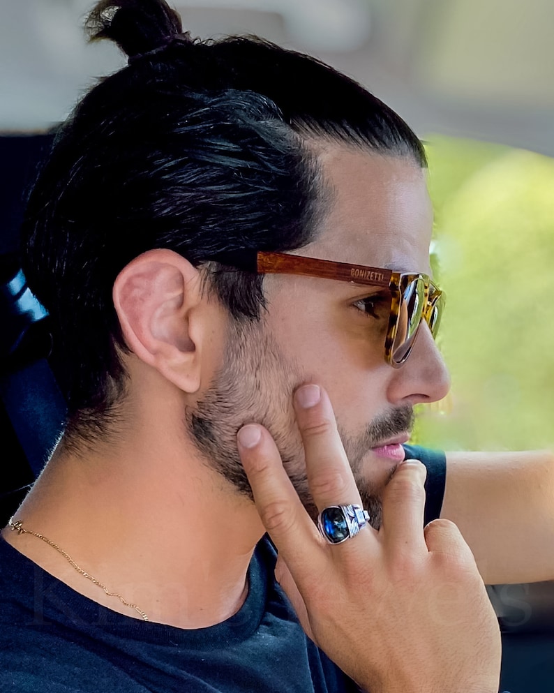 May include: A man with dark hair tied back in a bun is wearing brown tortoiseshell sunglasses with the brand name "DOMIZETTI" printed on the frame. He is wearing a blue shirt and a gold chain necklace. He is wearing a silver ring with a blue gemstone on his right hand.