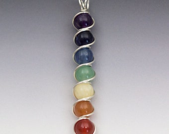 Puede incluir: Un colgante de alambre plateado envuelto con siete piedras de chakra. Las piedras son amatista, azul oscuro, azul claro, verde, amarillo, naranja y rojo.