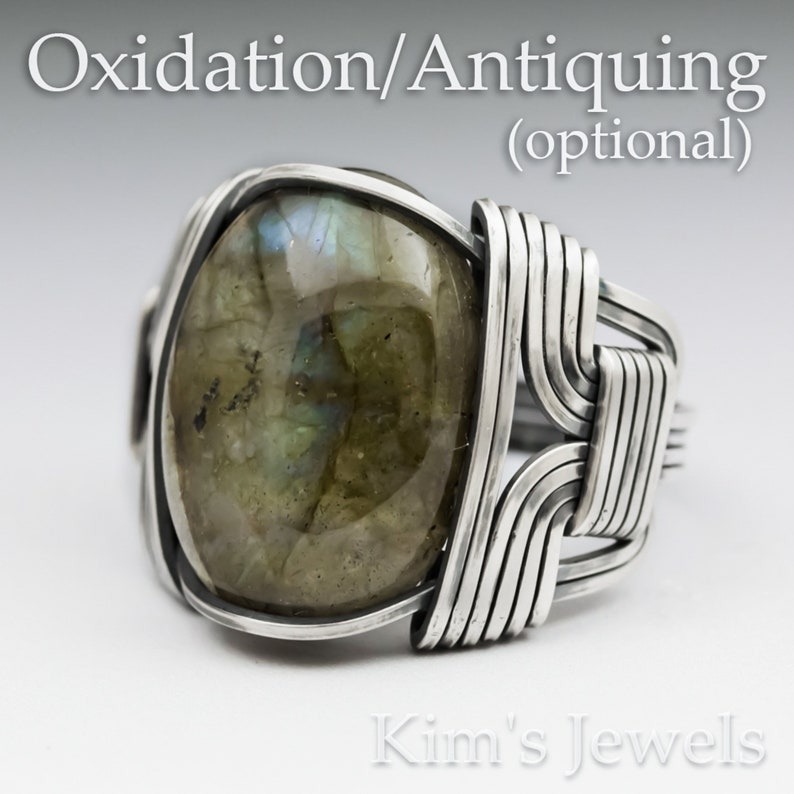 May include: A silver ring with a large, oval-shaped labradorite gemstone. The ring is made with wire wrapped around the stone, creating a unique and intricate design. The text "Oxidation/Antiquing (optional)" is visible above the ring.