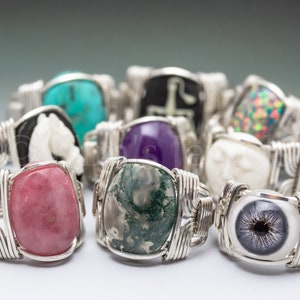May include: A collection of rings with various gemstone inlays, including a pink stone, a green and white stone, a purple stone, a turquoise stone, a white stone, and a black and white eye design. The rings are made with silver wire.
