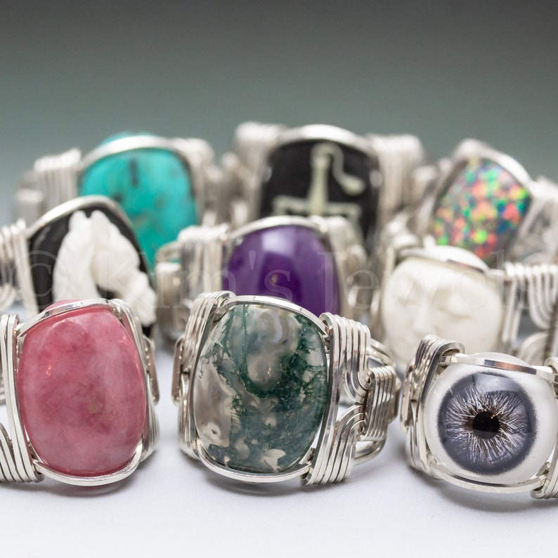 May include: A collection of rings with various gemstone inlays, including a pink stone, a green and white stone, a purple stone, a turquoise stone, a white stone, and a black and white eye design. The rings are made with silver wire.