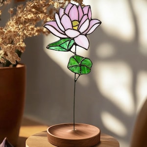 May include: A stained glass lotus flower with pink petals and green leaves on a wooden stand. The flower is on a wooden table with a brown vase and figs in the background.