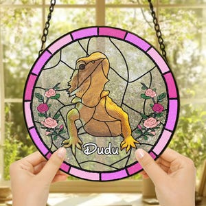Può includere: Una finestra rotonda in vetro colorato appesa con una cornice rosa e bianca. La finestra presenta un'immagine di un drago barbuto con il nome "Dudu" scritto sotto. Lo sfondo è un motivo floreale rosa e bianco.