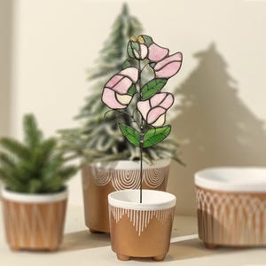 May include: A stained glass flower stake with pink petals and green leaves. The flower is in a small, gold-colored pot with a white rim and decorative lines. Other potted plants are in the background.