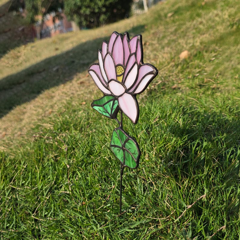 May include: A pink and white stained glass lotus flower with green leaves on a black metal stem. The flower is standing in a grassy area.