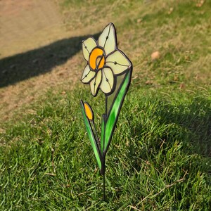 Puede incluir: Un narciso de vidrio tintado con p&eacute;talos amarillos y blancos y hojas verdes. La flor est&aacute; en una estaca de metal y se encuentra en un &aacute;rea cubierta de hierba.