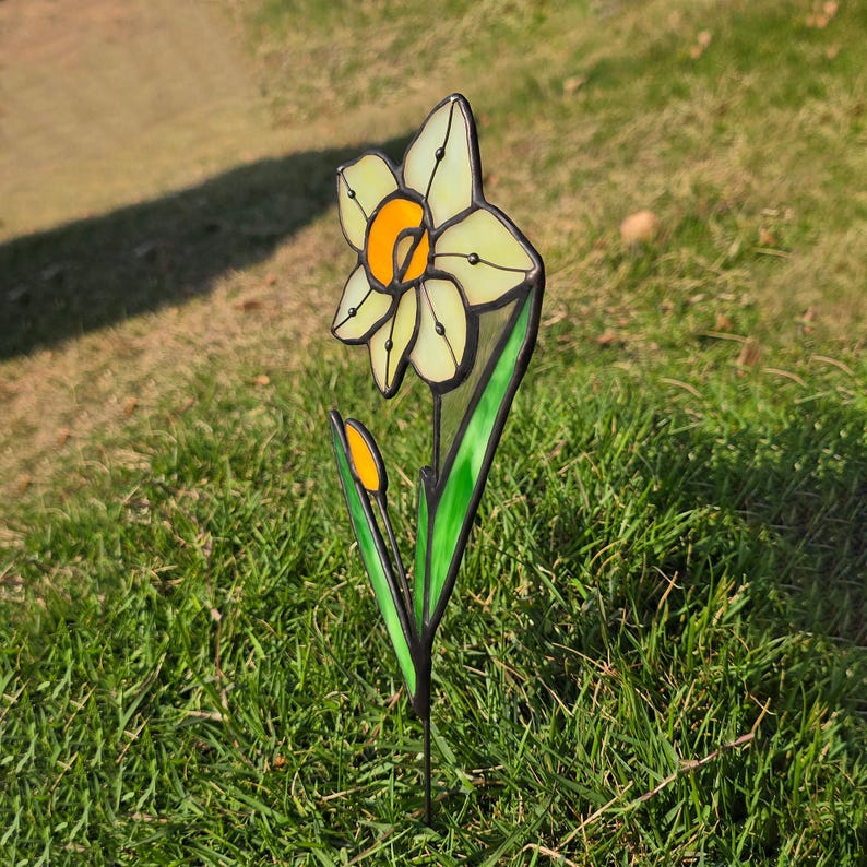 Puede incluir: Un narciso de vidrio tintado con p&eacute;talos amarillos y blancos y hojas verdes. La flor est&aacute; en una estaca de metal y se encuentra en un &aacute;rea cubierta de hierba.