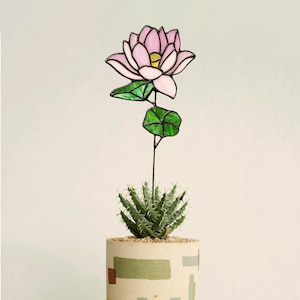 May include: A pink stained glass lotus flower with green leaves on a wire stem, placed in a pot with a succulent plant.