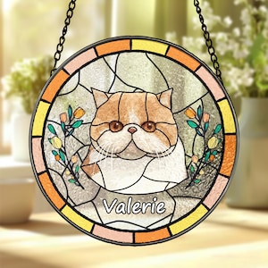 May include: A round stained glass window hanging from a chain. The window depicts a colourful cat with orange and white fur, surrounded by flowers and leaves. The cat's name, "Valerie", is written in a white cursive font at the bottom.