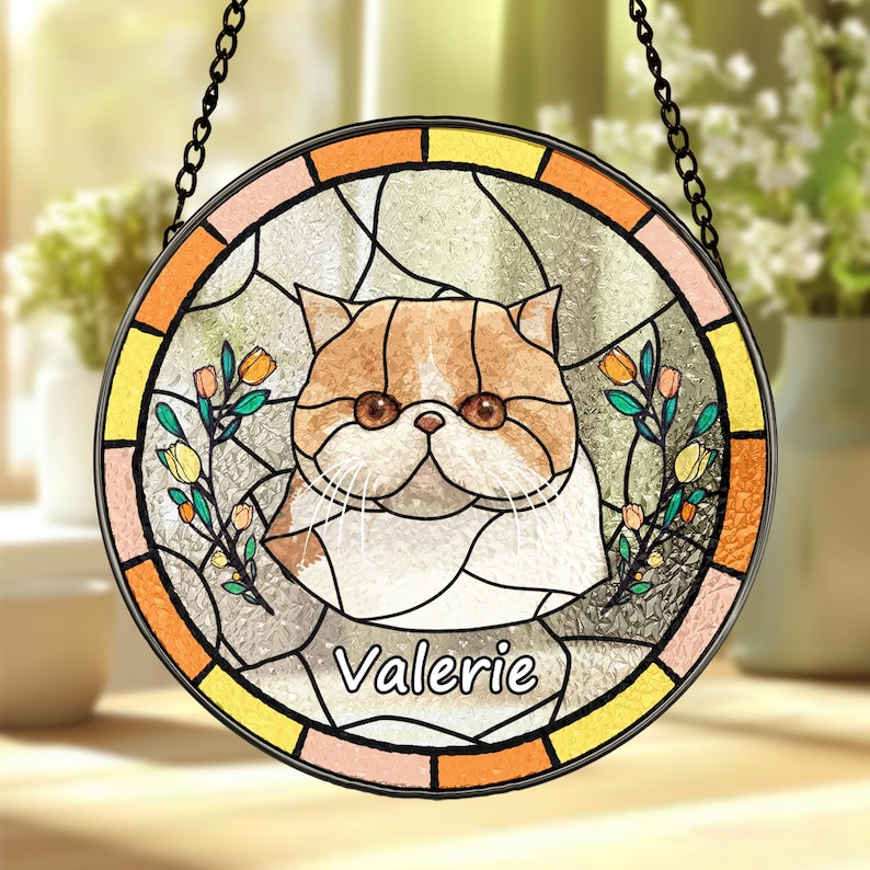 May include: A round stained glass window hanging from a chain. The window depicts a colourful cat with orange and white fur, surrounded by flowers and leaves. The cat's name, "Valerie", is written in a white cursive font at the bottom.