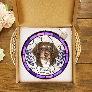 May include: A stained glass suncatcher featuring a brown and white dog with the name "Honey" in purple lettering. The suncatcher is in a purple and pink frame and is packaged in a brown box.