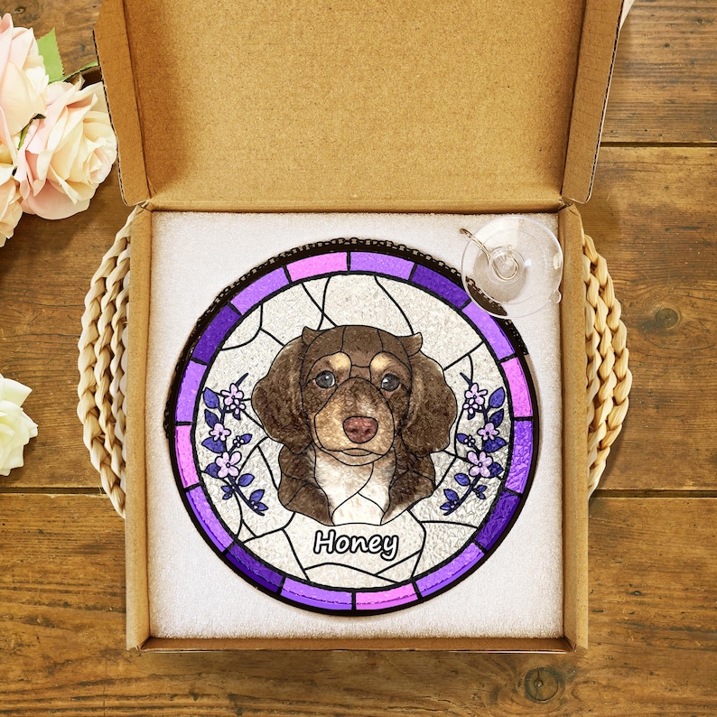 May include: A stained glass suncatcher featuring a brown and white dog with the name "Honey" in purple lettering. The suncatcher is in a purple and pink frame and is packaged in a brown box.