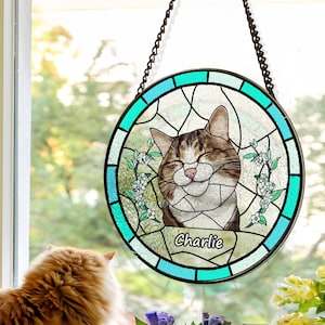 May include: A round stained glass window depicting a cat with brown and white fur. The cat is smiling with its eyes closed. The window is bordered in blue and green and has the name "Charlie" inscribed at the bottom.