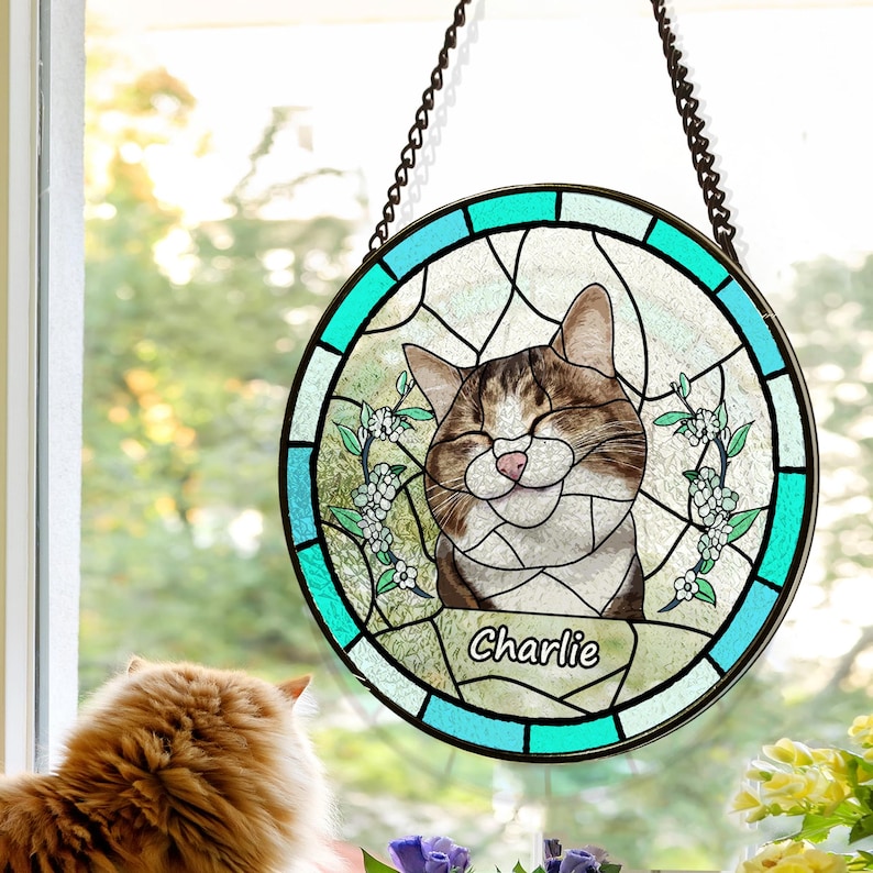 May include: A round stained glass window depicting a cat with brown and white fur. The cat is smiling with its eyes closed. The window is bordered in blue and green and has the name "Charlie" inscribed at the bottom.