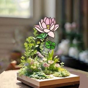 May include: A stained glass pink lotus flower with green leaves is displayed in a miniature garden setting. The garden is surrounded by a wooden frame and features a variety of green plants and small white flowers.