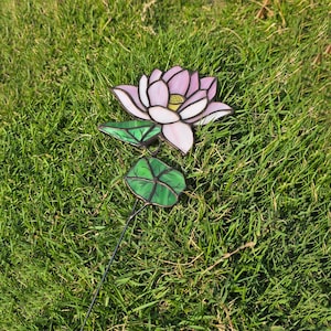 May include: A pink and white stained glass lotus flower with green leaves on a black metal stem. The flower is sitting on a bed of green grass.