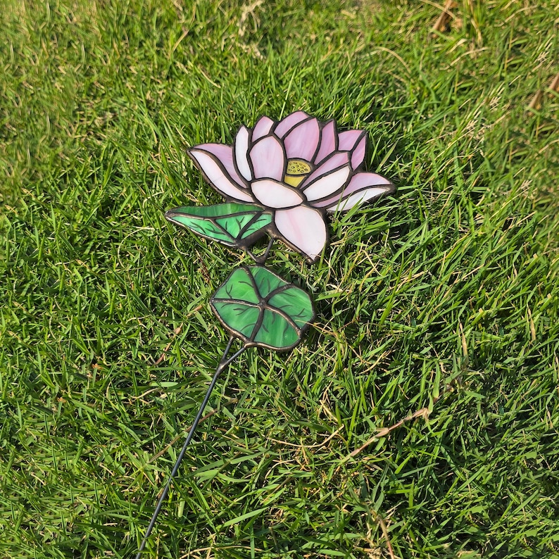 May include: A pink and white stained glass lotus flower with green leaves on a black metal stem. The flower is sitting on a bed of green grass.