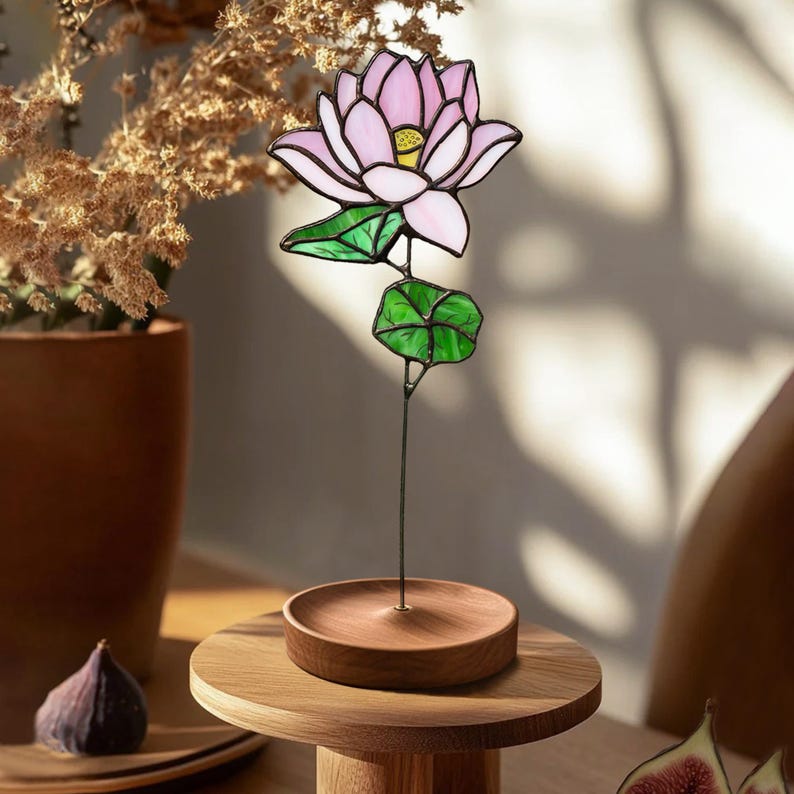 May include: A stained glass lotus flower with pink petals and green leaves on a wooden stand. The flower is on a wooden table with a brown vase and figs in the background.