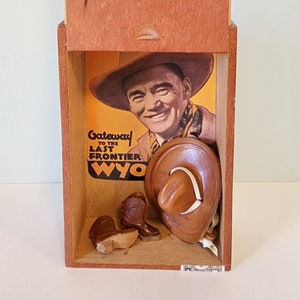 Könnte beinhalten: Eine Holzkiste mit dem Text "THE GRIFFIN'S" und "Toro Fuerte" auf der Vorderseite. Im Inneren befindet sich eine Vintage-Cowboy-Ausstellung mit einem Foto, Miniaturstiefeln und einem Hut. Auf dem Foto steht "Gateway to the Last Frontier Wyo."
