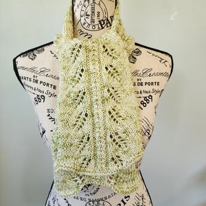 May include: A green and white knitted scarf with a lacy pattern. The scarf is draped over a mannequin with a vintage Parisian design.