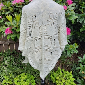 May include: Cream-colored, open-weave shawl with a geometric design. The shawl is displayed on a mannequin, showcasing its intricate knit pattern and scalloped edges. The background features vibrant pink flowers and lush greenery.