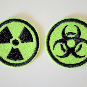 May include: Two round, embroidered patches. One features a black radiation symbol on a neon green background, the other a black biohazard symbol on a neon green background. Both have a black border.