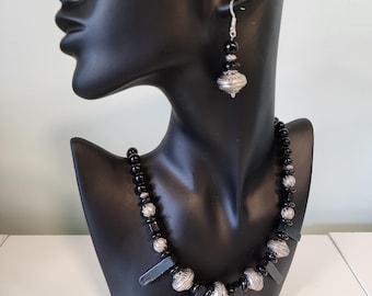 May include: A black and silver beaded necklace with long, black, rectangular pendant charms. The necklace is displayed on a black mannequin bust. Matching black and silver dangle earrings are also shown.