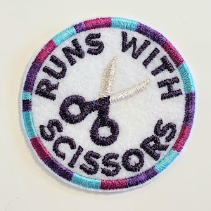 May include: A circular embroidered patch with a white background and a dark purple border featuring alternating segments of magenta and light blue. The patch displays a dark purple pair of scissors and the text RUNS WITH SCISSORS in dark purple.