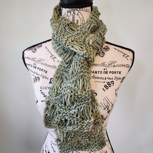 May include: A green knitted scarf with a textured pattern. The scarf is displayed on a mannequin with a vintage-style pattern.