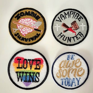 May include: Four embroidered iron-on patches. The first patch is gold with black text that says "Zombie Survival" and features a pink brain with a shovel and a pickaxe. The second patch is gray with black text that says "Vampire Hunter" and features a red cross, a bloody hammer, and a red cross. The third patch is white with black text that says "Love Wins" and features a rainbow heart. The fourth patch is white with blue text that says "Be Awesome Today" and features gold text.