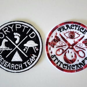 May include: Two embroidered patches. One black patch with white text reading "CRYPTID RESEARCH TEAM" around a crossed design with a Loch Ness Monster, footprint, and UFO. The other patch has red and white plaid with the words "PRACTICE STITCHCRAFT".