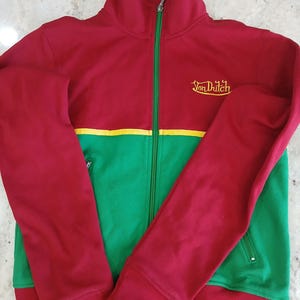 Puede incluir: Chaqueta deportiva roja, verde y amarilla con el texto "Von Dutch" bordado en el pecho. La chaqueta tiene cremallera completa, pu&ntilde;os acanalados y cuello alzado. Las mangas y la parte superior del cuerpo son rojas, mientras que la parte inferior es verde.