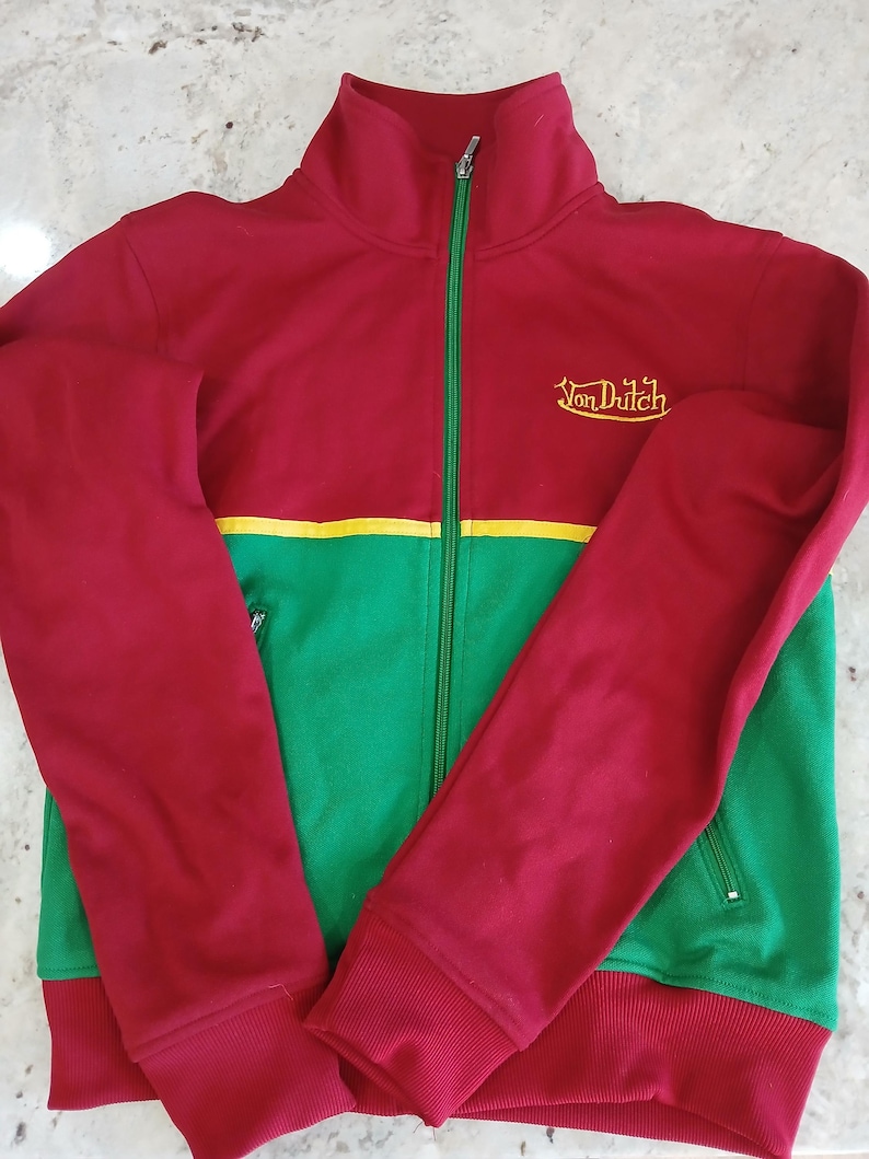 Puede incluir: Chaqueta deportiva roja, verde y amarilla con el texto "Von Dutch" bordado en el pecho. La chaqueta tiene cremallera completa, pu&ntilde;os acanalados y cuello alzado. Las mangas y la parte superior del cuerpo son rojas, mientras que la parte inferior es verde.